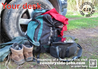 a pair of walking boots, a rucksack, rain jacket, ipad and camera bag on the ground near the front wheel of a car. Text reads: Your desk