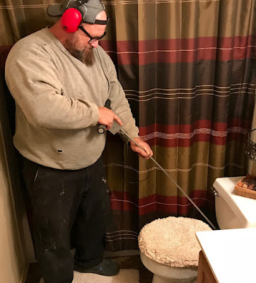 A plumber stands inside a bathroom wearing red headphones and holding a long pointy instrument. He is pointing the end of the instrument at the valve behind the toilet to listen for leaks