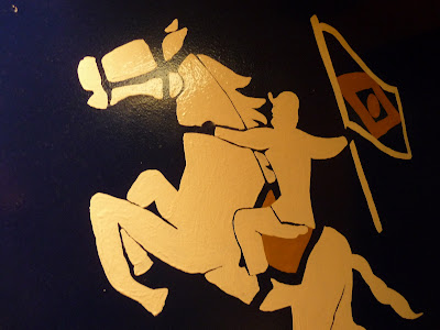 A simplistic painted mural of a man riding a horse into battle. He is carrying a flag with a diamond design.