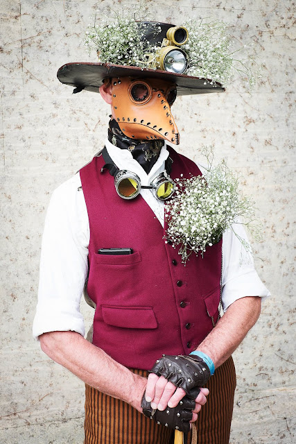 Steampunk Plague Doctor cosplay. Man wearing leather plague doctor mask, hat with babies breath flowers, vest, ascot, cane, and striped pants.