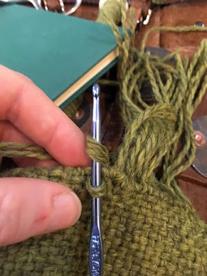 A white hand holds the edge of a piece of green cloth, through which a steel blue crochet hook is inserted, with a loop of light green yarn around the shaft of the hook, and the tails of the yarn wrapped over the shaft ready to be pulled through. At right, the cloth is fringed. In the background, a slim, unmarked, blue-green book is visible on rust-brown woven leather armour.