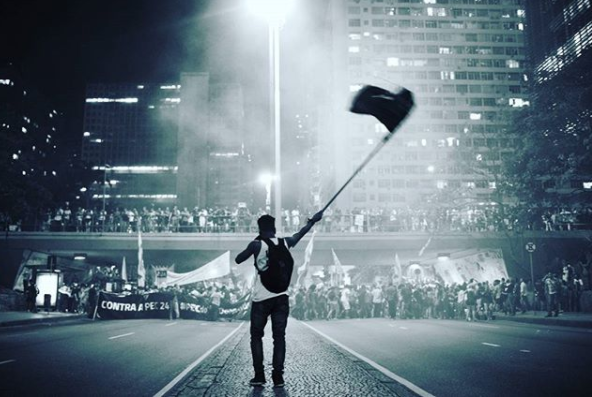 No meio da rua, jovem aparace no centro carregando uma bandeira, ele também carrega uma mochila nas costas. Mais a sua frente, uma multidão de manifestantes. 