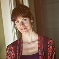 Photo of a woman grinning, wearing a purple blouse and long earrings, standing in front of a doorway 