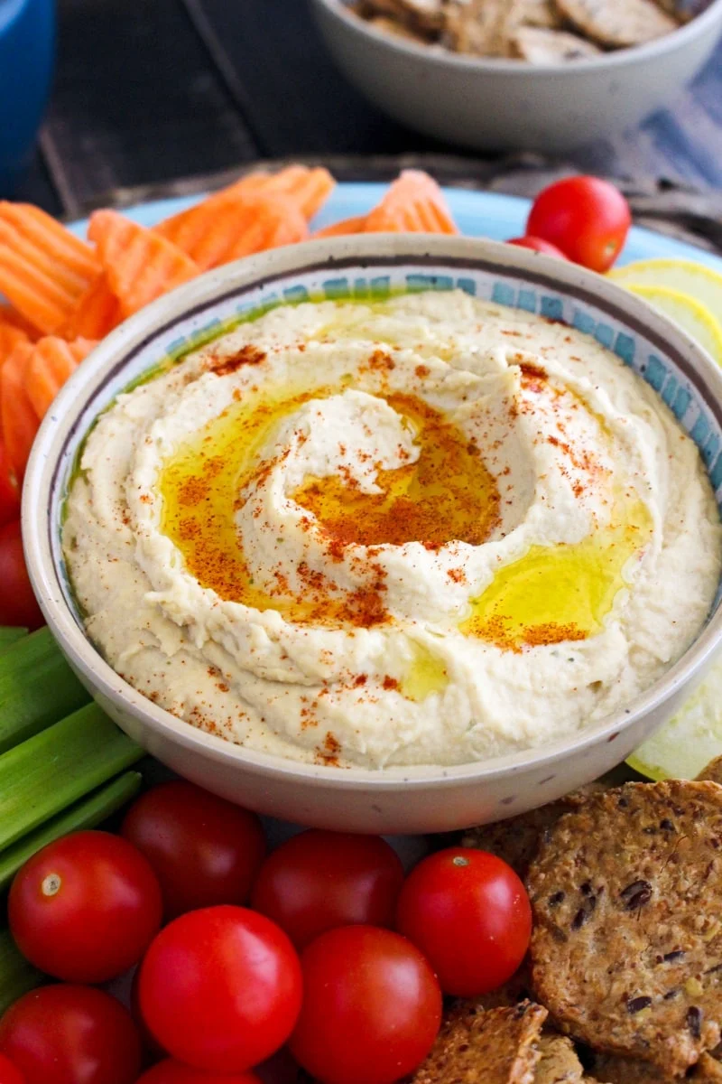 A bowl of hummus on a blue plate surrounded by crackers and chopped vegetables including cherry tomatoes, carrots, celery, and yellow squash on a dark wood table.