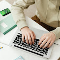 A close-up view of hands typing on a silver laptop keyboard, with the person wearing a cream-colored sweatshirt. Nearby on the white desk surface sits a green notebook and a smartphone with a glowing green screen, creating a clean, focused workspace environment for digital productivity.