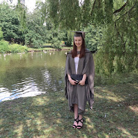 Pippa is stood in her grey coloured cap and gown by a small lake. She is surrounded by green trees, and there is wildlife in the lake. Pippa is smiling.