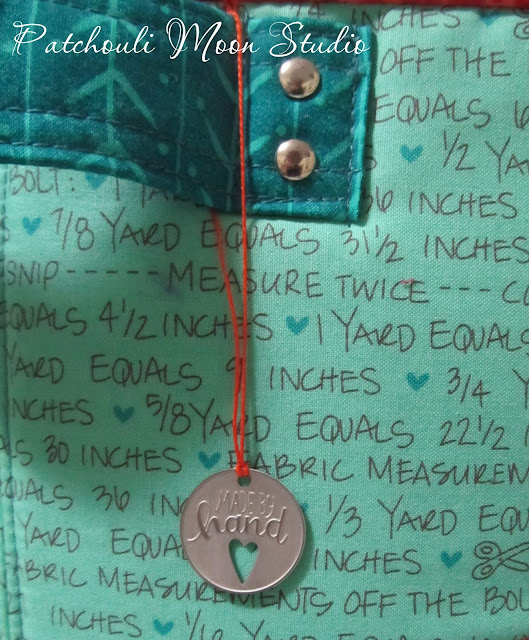 closeup of metal tag hanging from fabric container that says "made by hand" and depicts a cut out heart