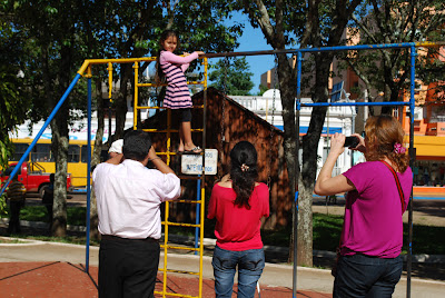 Os três aparecem de costas para a foto, com suas câmeras apontadas na direção da menina que está um pouco acima da altura deles, em um brinquedo de escalar. A menina está fazendo pose e olhando na direção do grupo.