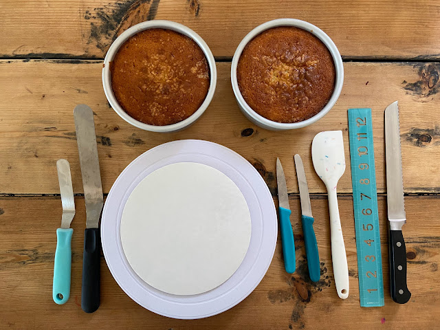 A photo of two small cakes in pans and lots of cake decorating tools, 2 offset spatulas, a turntable, two small knives, a spatula, a ruler, and a large serrated knife