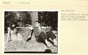 A photograph of a man and woman seated outside. Between them, a baby in a papoose is leaned against a tree trunk.