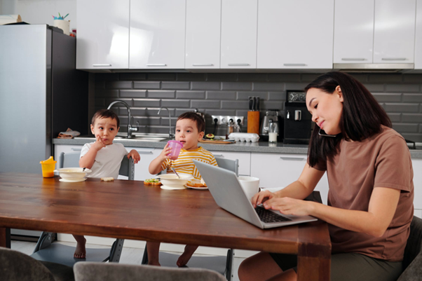 A mother works at a kitchen table as her children are eating breakfast.