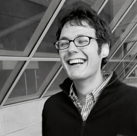 Ilya Kaminsky stands in an alcove with large windows. He is laughing and his eyes have closed. He has short, medium dark hair and wears glasses. He is wearing a striped button-down shirt and a dark pullover with a zipper.