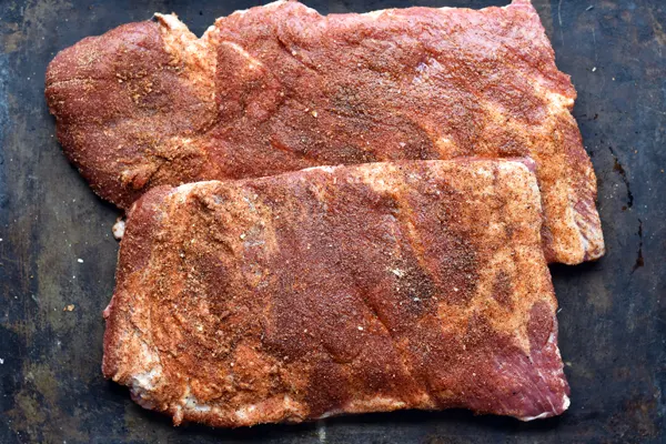 Raw St. Louis style ribs covered in a dry seasoning rub, ready to be slow cooked. Meat on a baking sheet, coated in a flavorful dry spice mixture, waiting to be cooked to perfection.