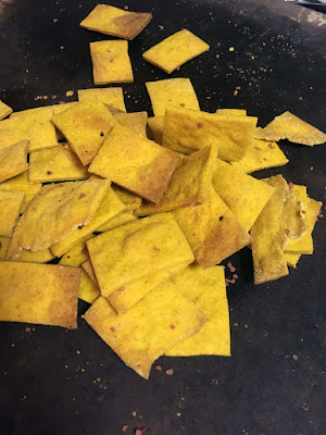 A pile of roughly rectangular bright yellow crackers collected on a black, slightly shiny pizza stone.