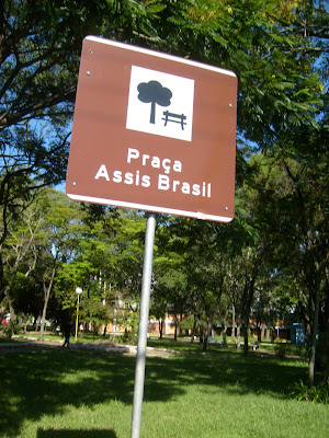 Placa de identificação com o nome da Praça Assis Brasil, mais conhecida como “Praça do Passo”. Ela é marrom e tem o desenho de uma árvore e de um banco.