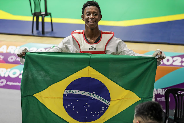 Henrique posa para foto segurando bandeira do Brasil. Ele é um rapaz negro, vestindo um quimono de taekwondo com colete vermelho