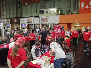 A load of people playing prototype games on tables with other people looking on. It's in a big hall and there are food concessions and toilets in the background