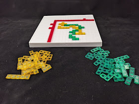 The same board as before, but with several rows and columns blocked off by a number of the red pieces to provide a smaller playing area, which is occupied by several of the yellow and green pieces, the unused of which are piled next to the board.