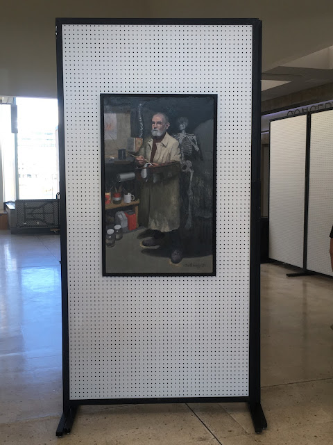 Oil painting of man with beard and glasses wearing tan raincoat and holding a paint brush and a wooden palette, hanging on a pegboard panel in gallery.