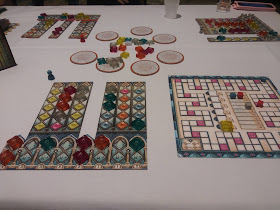 A game of Azul: Stained Glass of Sintra in progress. Several player boards with translucent coloured tiles arranged on them. Seven round discs, one with four of the coloured tiles on it, sit in a ring in the centre of the table, with a number of the coloured tiles in the centre of the circle. There is a score tracker nearby, with coloured scoring markers on it.