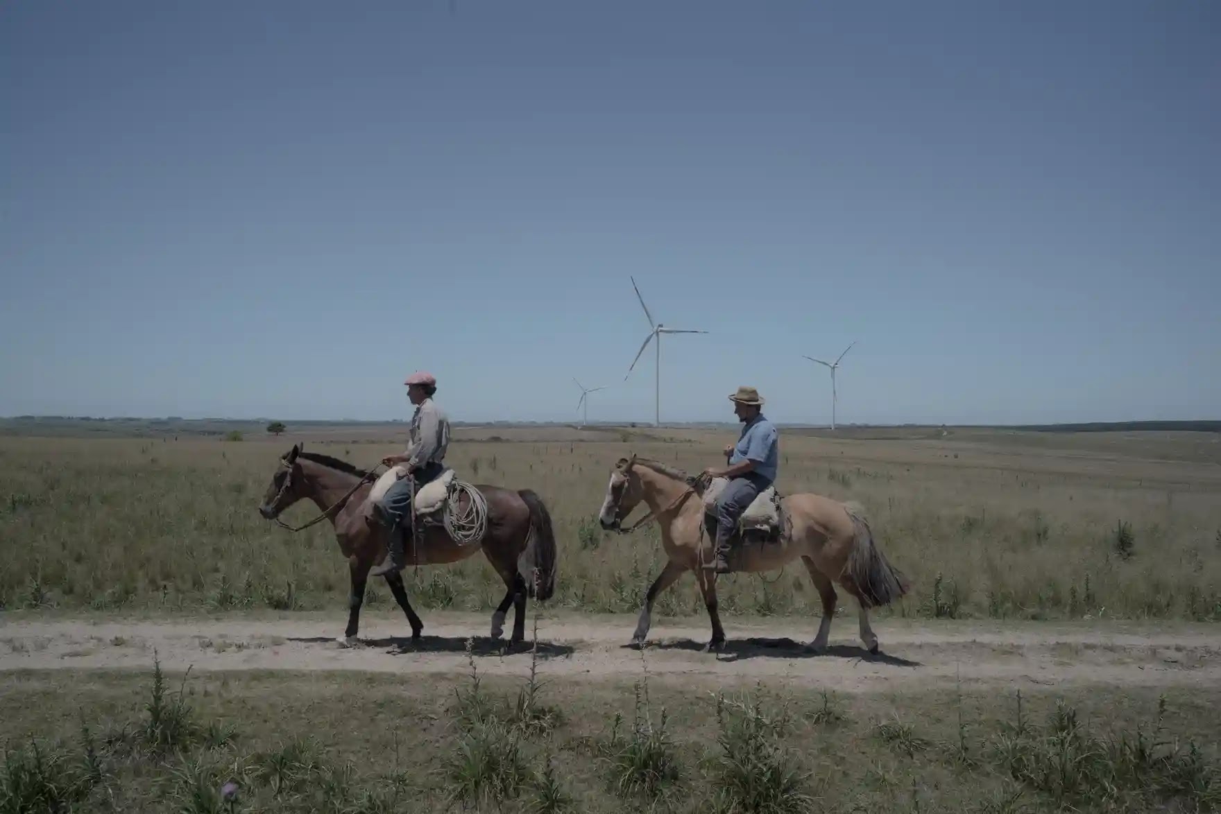 Two men ride horses along a track on a plain, in the background are several wind turbines