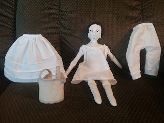 The doll from above, wearing only a white cotton chemise; beside her are a white petticoat with three tucks, a pair of long white drawers with tucks at the hem, and a set of cream-colored linen stays.