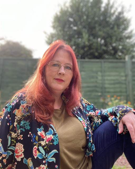 A redheaded white woman sits on the ground. She is wearing an olive top and navy jeans, with a loose, kimono style top over it all. The top is navy and printed with an Asian-inspired pattern of peonies and chaffinches in turquoise, pink, olive and cream on a navy background.