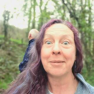 Lisa Southard pulling a What Have We Started face at the camera- Mr wanders in the background, in the scrubby woods, edged by trees in bud