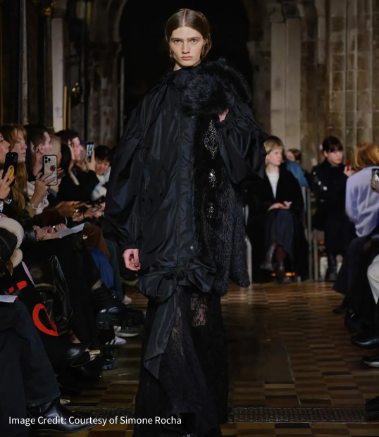 Photo of female model walking for Simone Rocha at London Fashion Week 2024. Model is Wearing a floor length black lace gown with puffy black overcoat. Both dress and coat is baggy in style giving it a contemporary feel