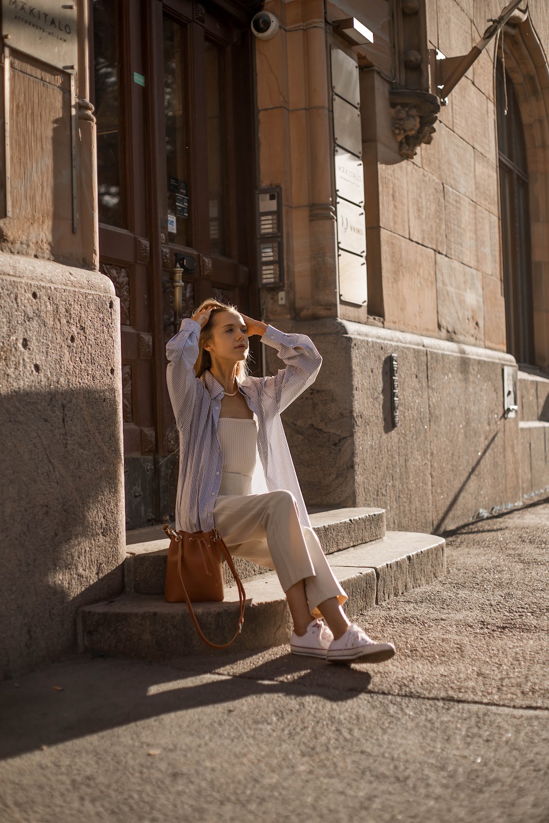 Nainen istuu portailla auringossa päällään beiget housut, beige tuubitoppi ja sinivalkoraidallinen kauluspaita