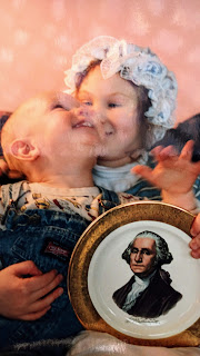 A little girl in a colonial bonnet and a littler boy in overalls snuggle, holding a gilt-edged plate with a portrait of George Washington