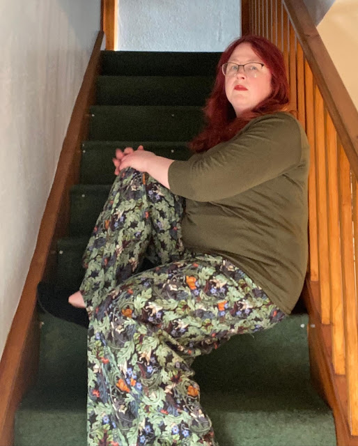 A redheaded white woman sitting on some stairs. She is wearing a close-fitting olive-green long-sleeved top and baggy linen trousers with a pattern of swirling green leaves on a black background, interspersed with brightly-coloured flowers.