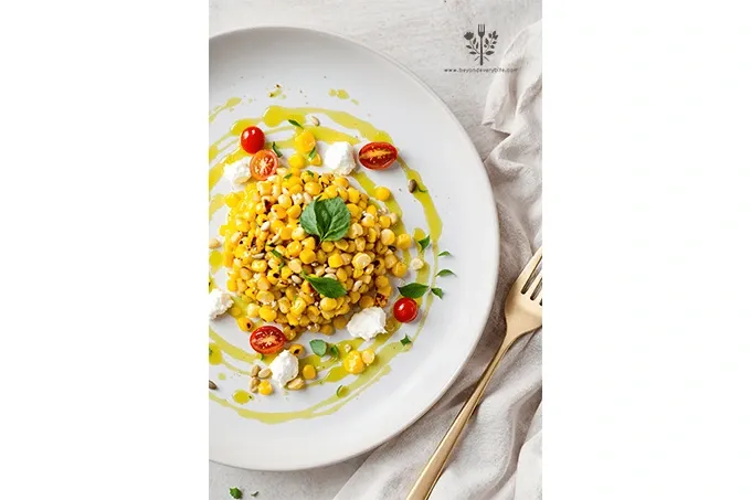 Charred Corn Salad with Basil Oil plated with cherry tomatoes, goat cheese, toasted pine nuts, and microgreens on a white ceramic plate.