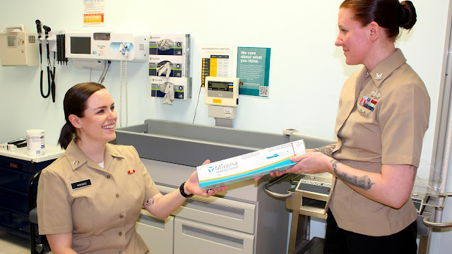 Two women in tan military uniforms are smiling and holding a long, narrow box labeled "Mirena" in a clinical setting. The environment includes medical equipment and informational posters, suggesting a healthcare facility within a military context.