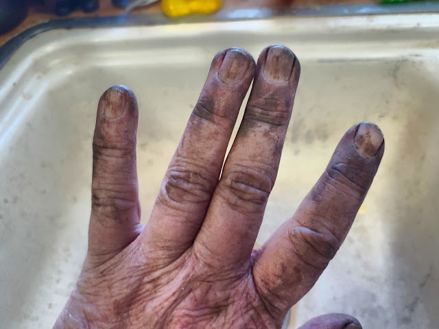 A hand is shown with dirt under the fingernails and covering the skin, suggesting recent gardening or manual labor. The fingers are spread apart, and the background features a blurred sink.