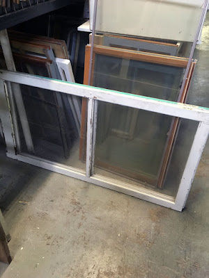 A number of salvage windows stacked on edge on a cement floor, under a metal shelf. Some small, wood-framed windows are pulled out and leaning against the side of a white shelving unit, and a double-sashed, white-framed window rests on its side at the front of the stack.