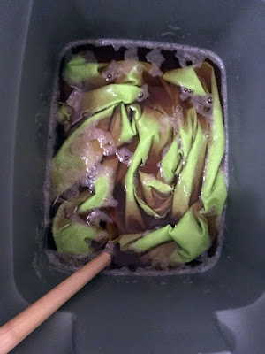 A photo from above of bright green fabric submerged in thin brown tea in a grey tote, with a dowel sitting in the lower left corner of the tote.