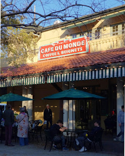 The exterior of a cafe, folks seated under green umbrellas, a red and white sign overhead marks is as 'Cafe du Monde, Coffee & Beignets'.