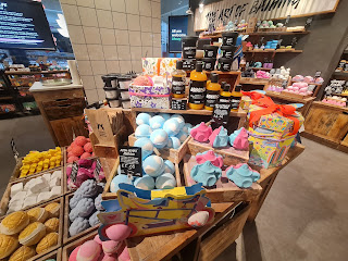 A wide shot photo of a selection of light and brightly coloured spherical bath bombs, cylindrical clear bottles filled with orange liquid with mimosa on a back label and other products in light brown shelves and square light brown boxes on a light brown table on a bright background