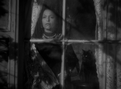 A stern-looking female housekeeper stares out of a window, accompanied by a black cat with gleaming eyes