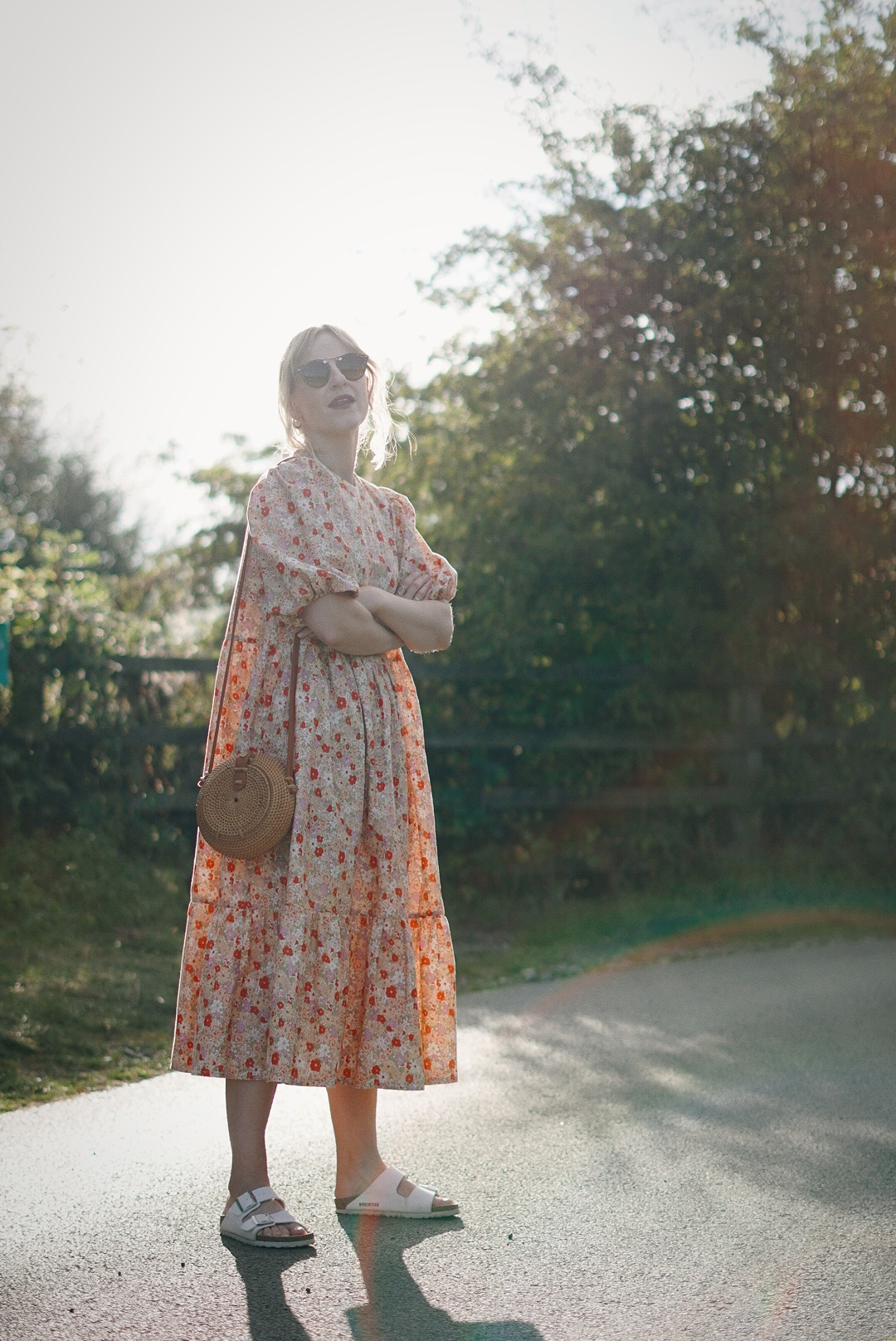 Full length photo of Amy in a peach coloured floral oversized maxi dress. The sun is behind her creating a haze through the trees.