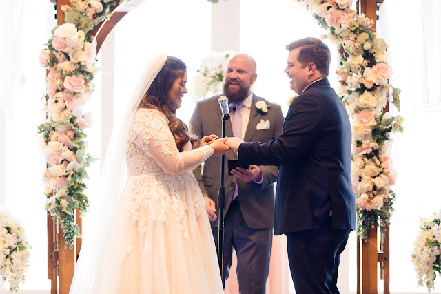 May 1st is a special day in the lives of Jonathan and Katelin, for it was on this day one year ago that he proposed at the Myriad Gardens in Oklahoma City.  Both Katelin and Jonathan grew up in Stillwater, went to the same middle and high school, but didn’t actually get to know each other until one of Katelin’s friends suggested that she invite Jonathan to the Senior Prom.  Katelin didn’t think that was a good idea at all, since she described him as “a very shy, nerdy kid,” and she was a “social butterfly.”  Little did she know that “we would really hit it off…and things have just gotten better every day since!”     And what was not to love about this day?  The bride and groom chose the Sparrow in Stillwater as their venue, with a rustic, architecturally modern barn, and 30 beautiful acres to roam in.   Katelin and her bridesmaids spent the morning, tucked away in their own getting-ready suite, finishing up with hairdresser, make-up artist, and assisting the bride into her gorgeous gown.  They were also trying to be a calming influence, but as the time for the First Look arrived, everyone was excited…and emotional.  Especially the newlyweds-to-be!     Katelin and Jonathan exchanged sweet letters during the First Look, which brought joyful tears to each of them, and then they were whisked off to create some magical couples’ portraits.  Wedding party pictures followed, as the bridesmaids gathered around Katelin, and the groomsmen’s personalities emerged as they enacted the “Mario” jump!    At last the hour came for the ceremony to begin.  The bridesmaids, dressed in pastel shades of pink and lavender, came down the staircase on the hand of their respective groomsman, while Jonathan waited patiently at the altar for his bride.  Added to the wedding party were the couple’s two Golden Retrievers—the “flower girl and ring bearer.”  The bride was stunning in her lace-embroidered, elegant wedding gown, carrying a lush floral bouquet consisting of white roses, white ranunculus, and lush greenery.     After the couple exchanged vows, guests were invited to participate in a buffet dinner while they enjoyed the festivities of the evening…the first dance, dances with the respective parents, toasts to the bride and groom, and dancing.  The culmination of the evening took place when Jonathan, in light-hearted fun, smashed some of the wedding cake on Katelin’s face, only to be sprayed with champagne when the bottle popped off as he was opening it!  True to her humorous good nature, Katelin took it all in stride!     This is one of those weddings we will remember, reminding us that soul mates do find one another in the most wonderful of unexpected ways…and we wish this delightful couple a lifetime of happiness together!