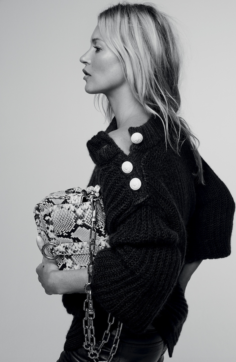 Woman with long straight blonde hair with highlights and middle part wearing black leather pants and a large knit oversized sweater with large gold buttons on shoulder partially open with hand on hips holding square shoulder bag in brown python print with chain shoulder strap in front of a white background