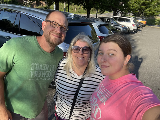 Dad, Me, and then Elizabeth taking a selfie in a restaurant parking lot. Dad is on the left, I am in the center, and Elizabeth is on the right hand side. Our arms are around each other. Dad is wearing his glasses and his green fat-heads tshirt. I am wearing sunglasses with my hair down and straightened. I am wearing a black and white striped t-shirt with my purse strap across my chest hanging over my left shoulder. Elizabeth is wearing her pink dr. pepper tshirt with her hair pulled back into a ponytail. The sky is blue and the sun is out. In the background of the photo are random cars parked in the restaurant parking lot.