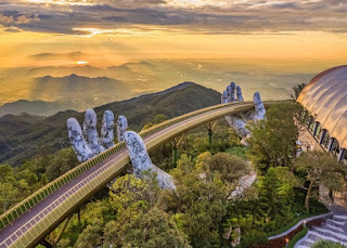 Foto. Apoiada por mãos gigantes em concreto, ponte a 1.400 metros de altura em uma área com vegetação é  atração no Vietnã. Ao longe, uma névoa paira sobre a paisagem ao entardecer.