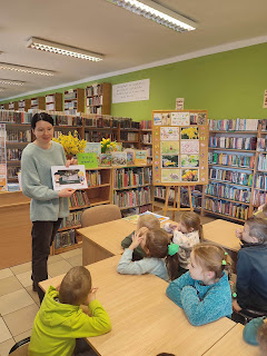 Pani bibliotekarka pokazuje dzieciom zwierzęta zwiastujące wiosnę. Tło: sala biblioteczna, regały z książkami.