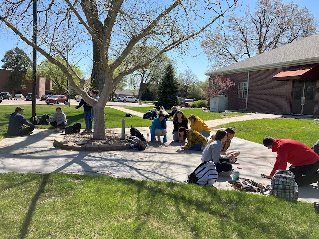 Outside, beautiful day. Young adults huddle under a tree in groups to create sidewalk chalk drawings.