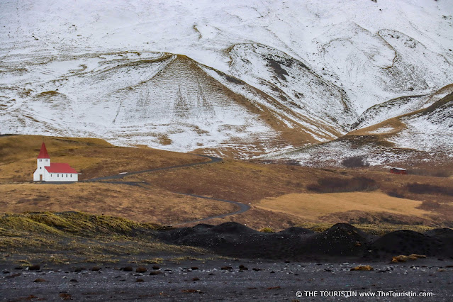 A white red-roofed church on a black lava beach in front of a green covered and partially snow-capped mountainscape.