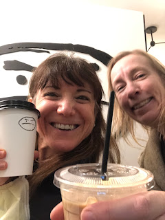 A selfie of me and Elaine holding coffee cups with a funny cartoon "grumpy" face on the wall behind us.