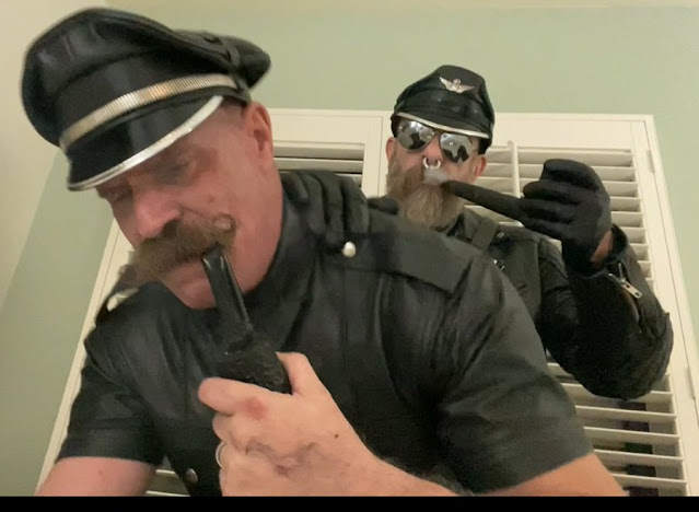 Two men wearing leather gear with mustaches smoking pipes and pounding each other's butts